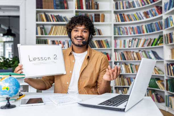 An enthusiastic online teacher giving a lesson from a home office with a whiteboard and a laptop in a bookshelf background.