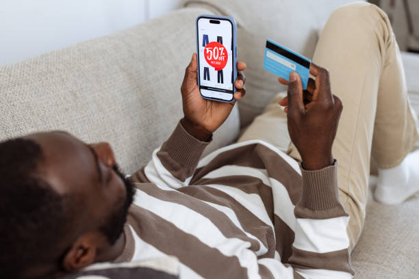 A man lies on a sofa using his smartphone and a credit card for online shopping, viewing a discount ad. The scene conveys relaxation, technology use, and digital commerce convenience.
